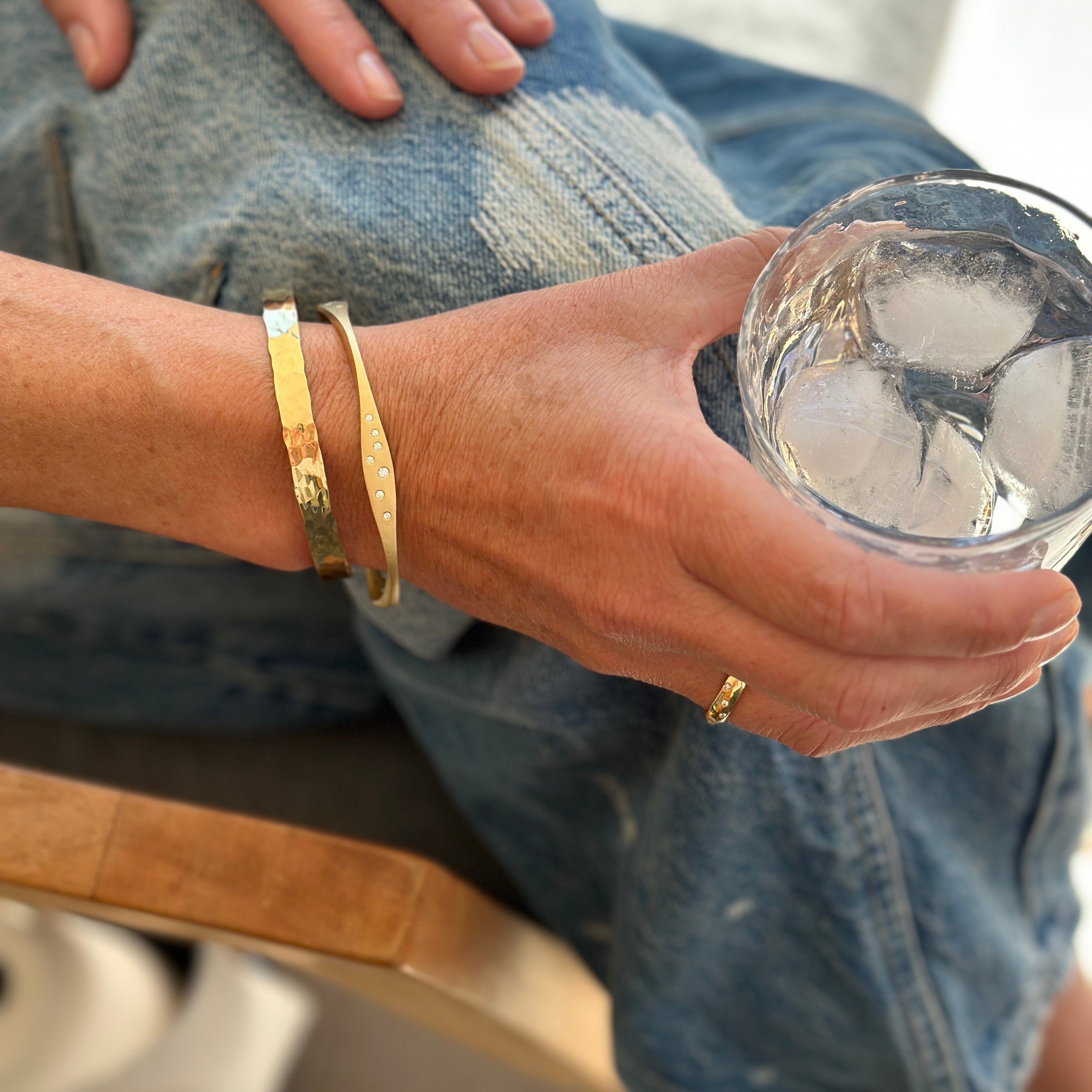 Person in blue jeans wearing gold bracelets and a gold ring, sitting and holding a glass of ice water.