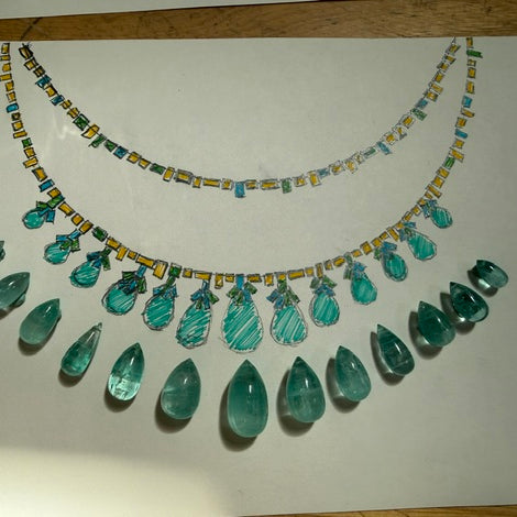 A hand-drawn or colored sketch of a statement necklace on a white sheet of paper, placed on a wooden surface. The necklace design features multiple rows of faceted teal and green teardrop gemstones with yellow and blue accents along the chain.