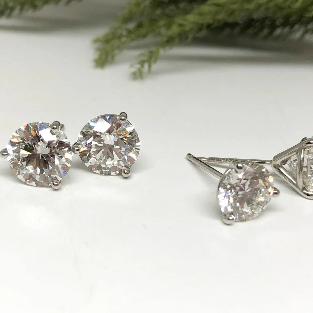 Pair of diamond stud earrings on a white surface with greenery in the background