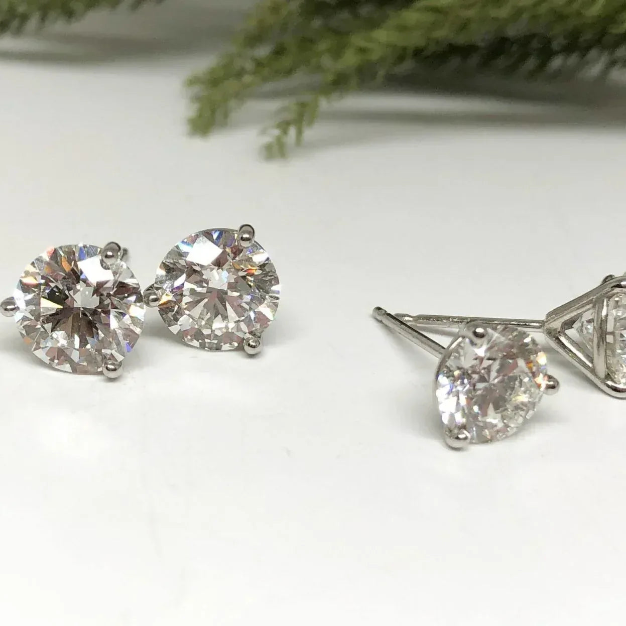 Pair of diamond stud earrings on a white surface with greenery in the background