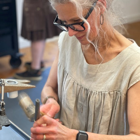 A woman with blonde hair, glasses, and a beige sleeveless top is shown from the waist up, working at a metalworking bench with a hammer and metal rod. Another person is blurred in the background.