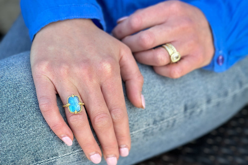 Woman in a blue shirt sitting with hands on jeans wearing a blue gemstone ring and gold ring