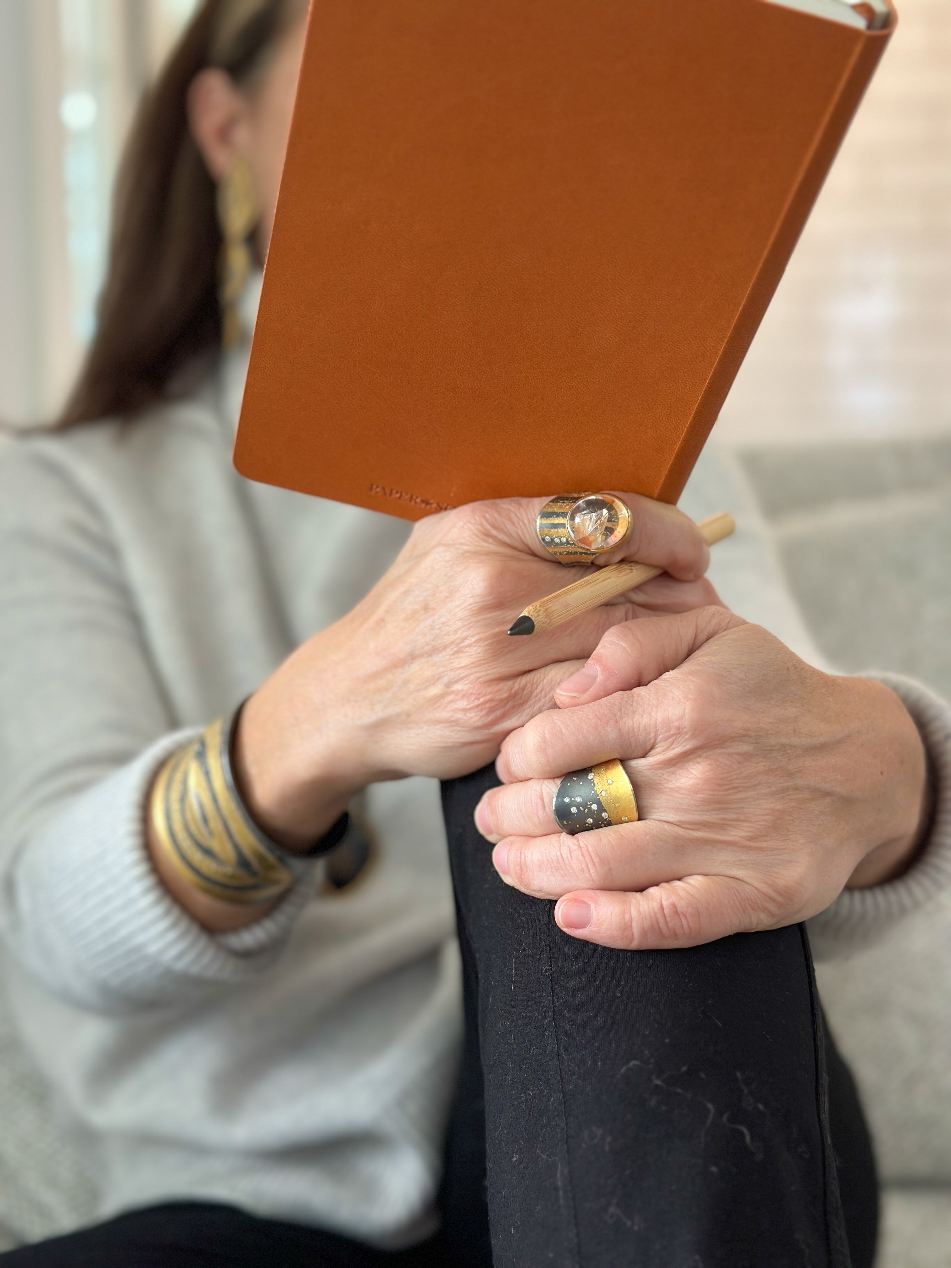 Woman reading a book with her hands on her knee wearing Gold & Black jewelry