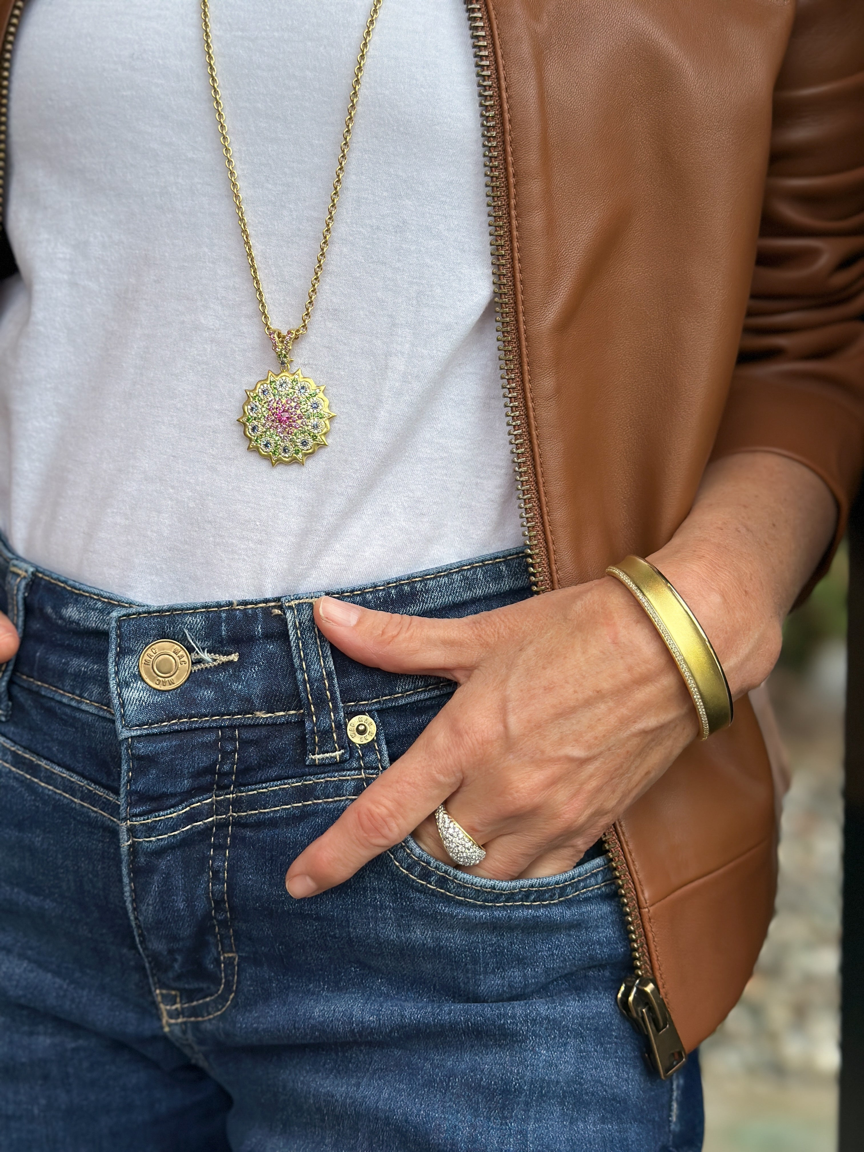 Woman wearing jeans, a brown leather jacket and white shirt with her hand in her pocket.  She is wearing long gold necklace with a colorful pendant, gold ring and gold bracelet