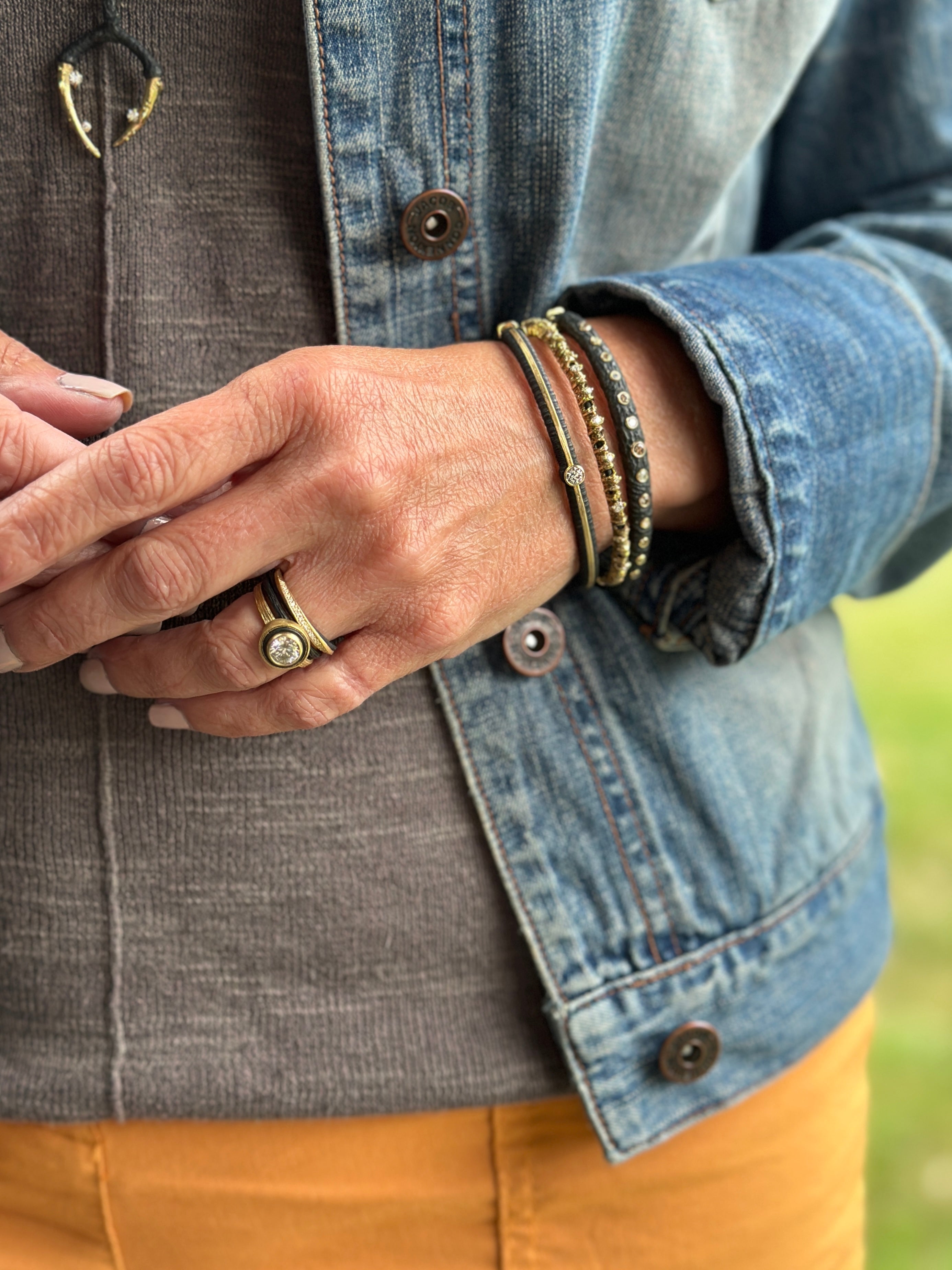 Person wearing brown bottoms with a denim jacket over a grey shirt wearing Sarah Graham black, gold & gold bracelets, rings and pendants with diamonds.