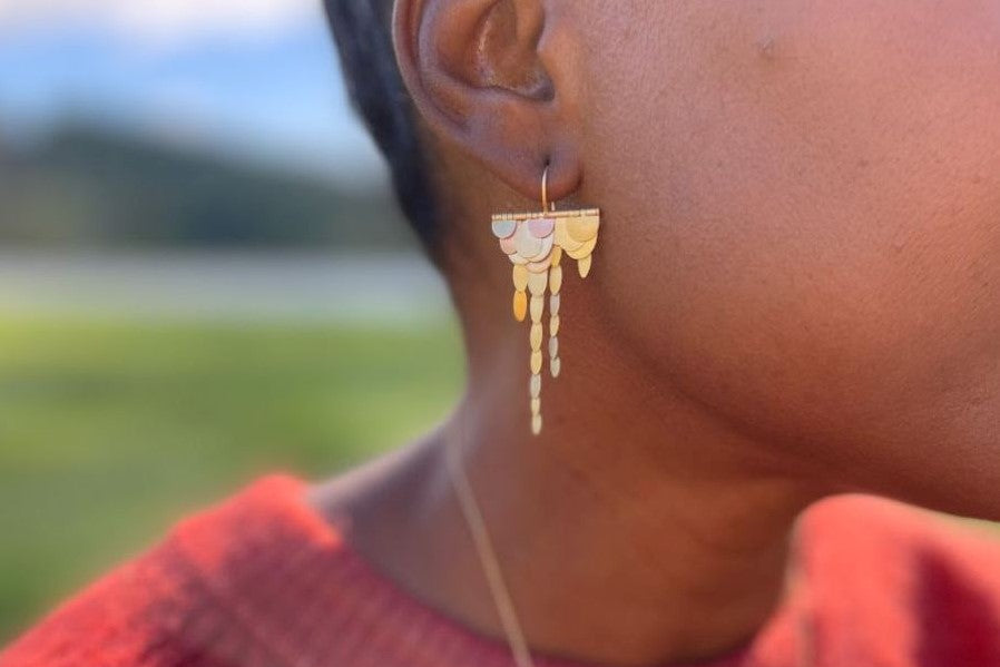 Woman in a red shirt wearing Sia Taylor dangle gold and white gold earrings and a gold necklace with grass and mountains in the background.