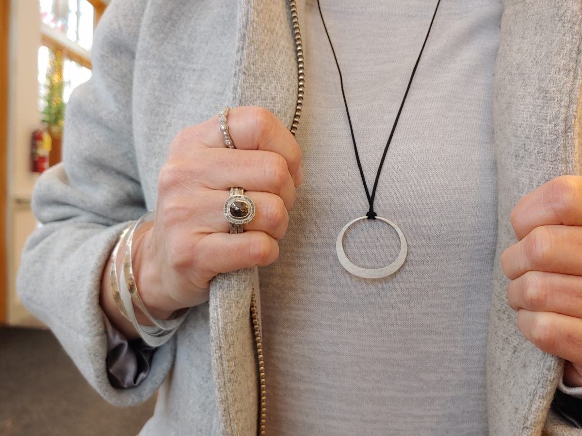 Man in grey wearing Toby Pomeroy Charm on a black cord necklace, two silver rings and bracelets