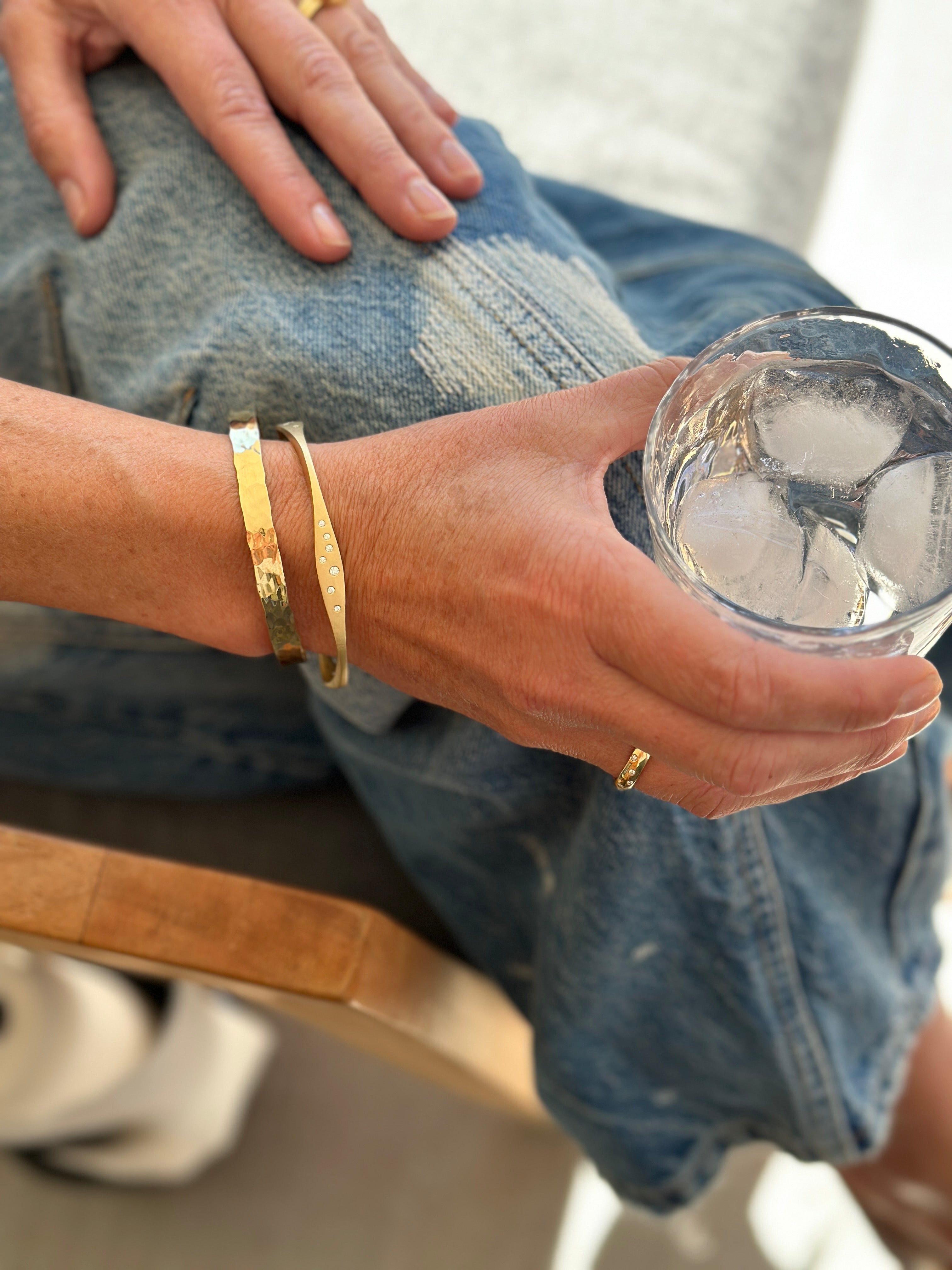 Person in blue jeans wearing gold bracelets and a gold ring, sitting and holding a glass of ice water.