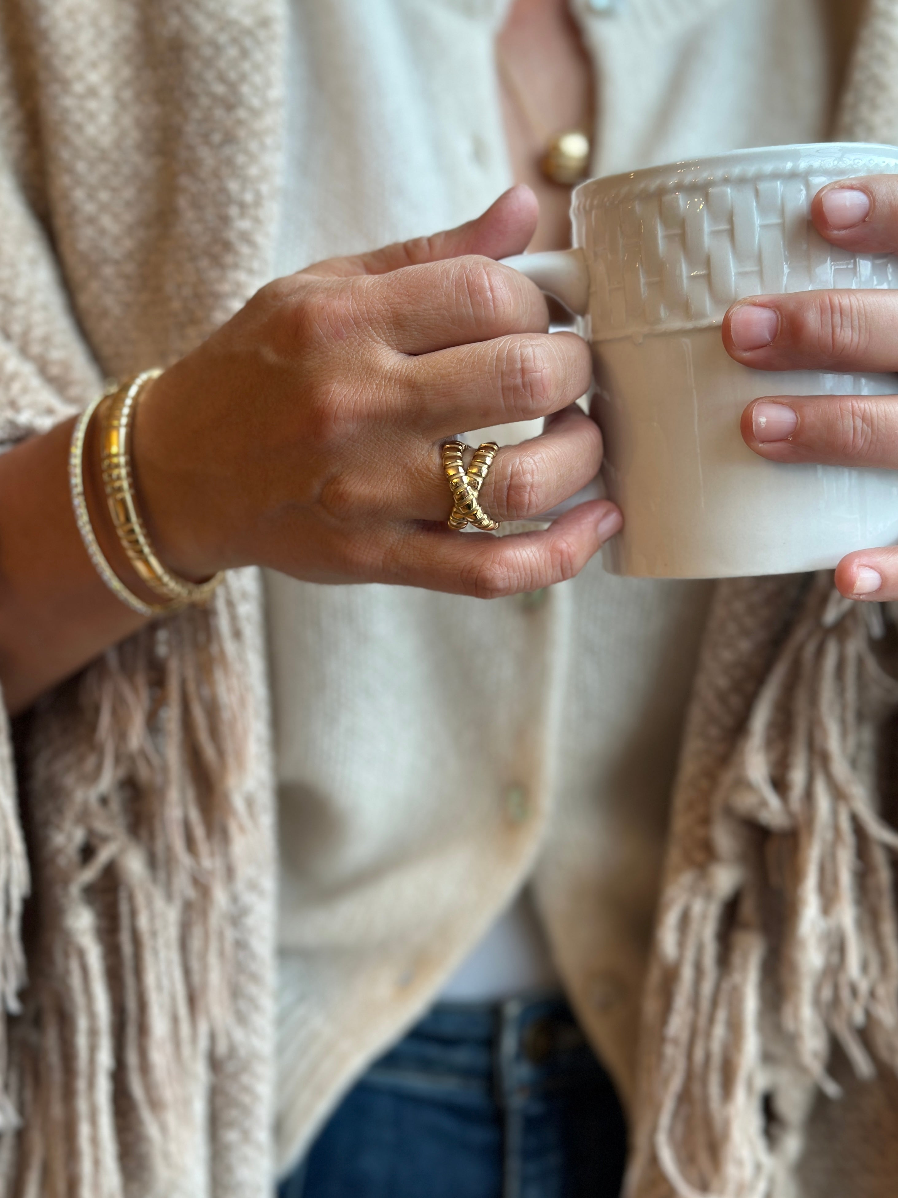 Woman in beige top wearing gold and diamond bracelets and a gold ring drinking coffee out of a white mug