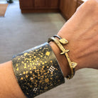 A hand wears two bracelets: a wide Sterling Silver Yellow Gold and Diamond Cuff, and a thinner gold bracelet with three small, irregular stones. The softly blurred background shows indoor flooring and cabinets.