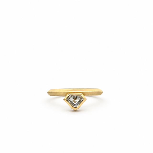 A gold ring featuring a flat, elongated bar with a small, faceted diamond-shaped bezel holding a clear gemstone, set against a plain white background.