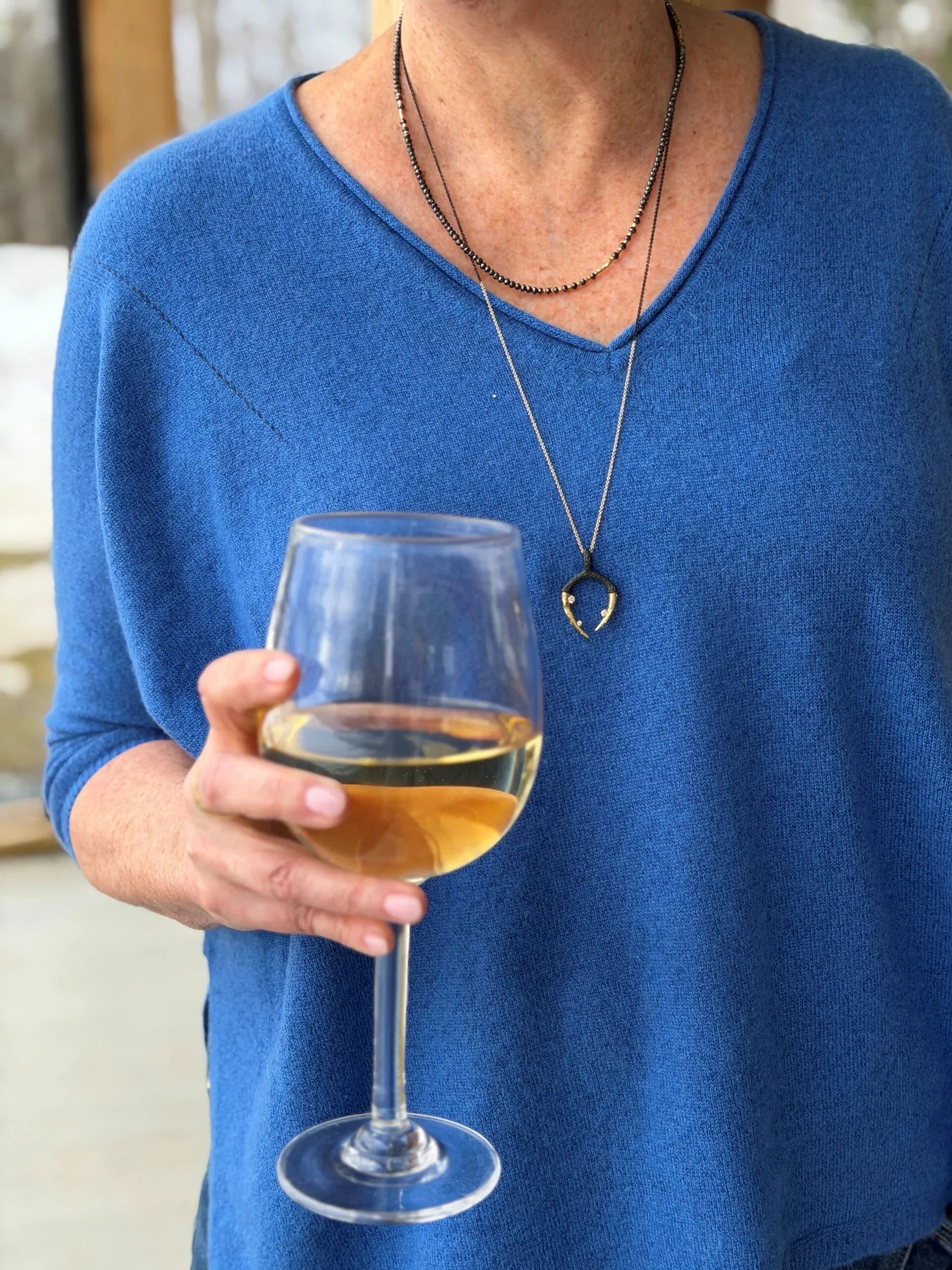 Person holding a glass of white wine wearing a blue shirt and necklace with a heart pendant.