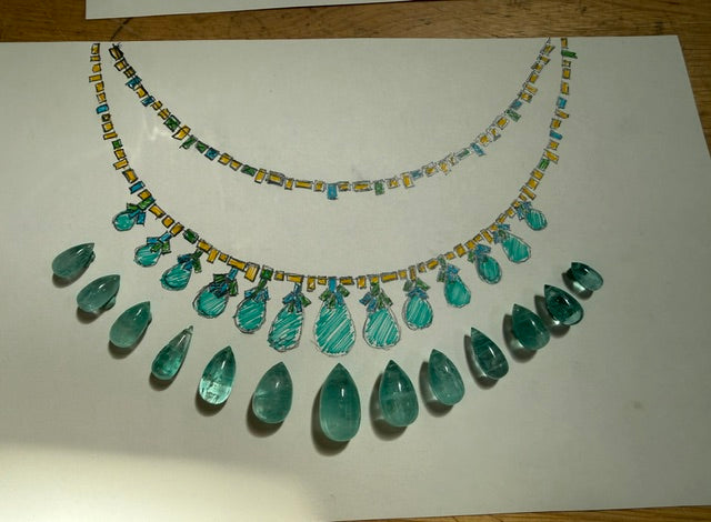 A hand-drawn or colored sketch of a statement necklace on a white sheet of paper, placed on a wooden surface. The necklace design features multiple rows of faceted teal and green teardrop gemstones with yellow and blue accents along the chain.