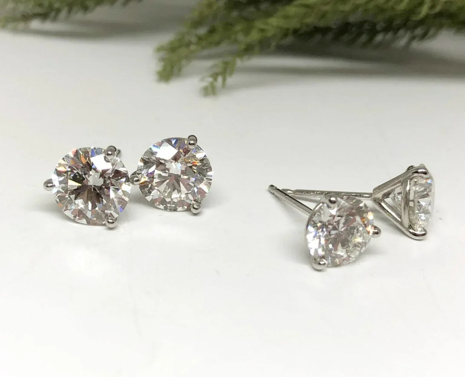 Pair of diamond stud earrings on a white surface with greenery in the background
