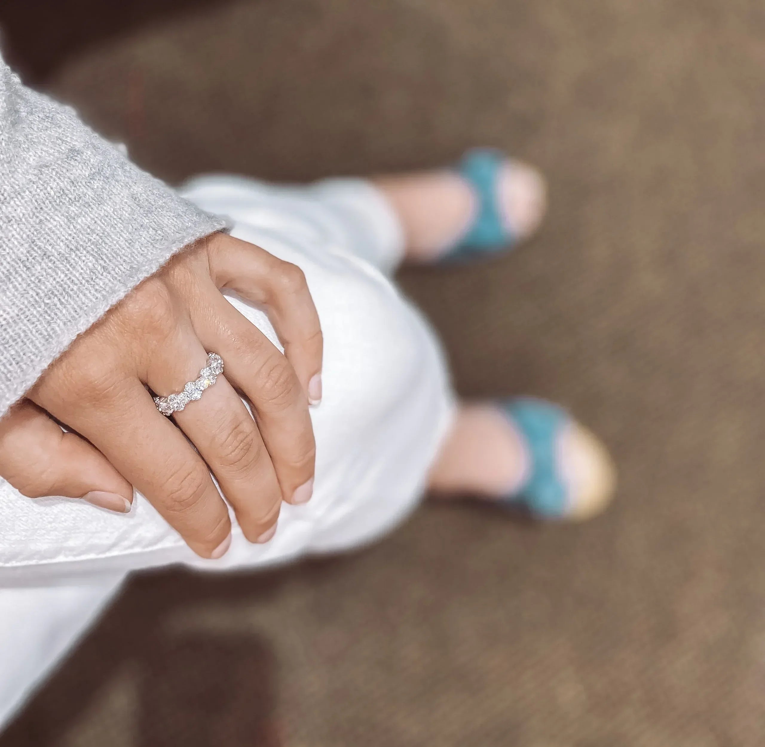 A person wears a White Gold and Diamond Eternity Band on their left hand, resting it on white pants. They also wear a grey sweater and teal sandals, set against a brownish floor background.