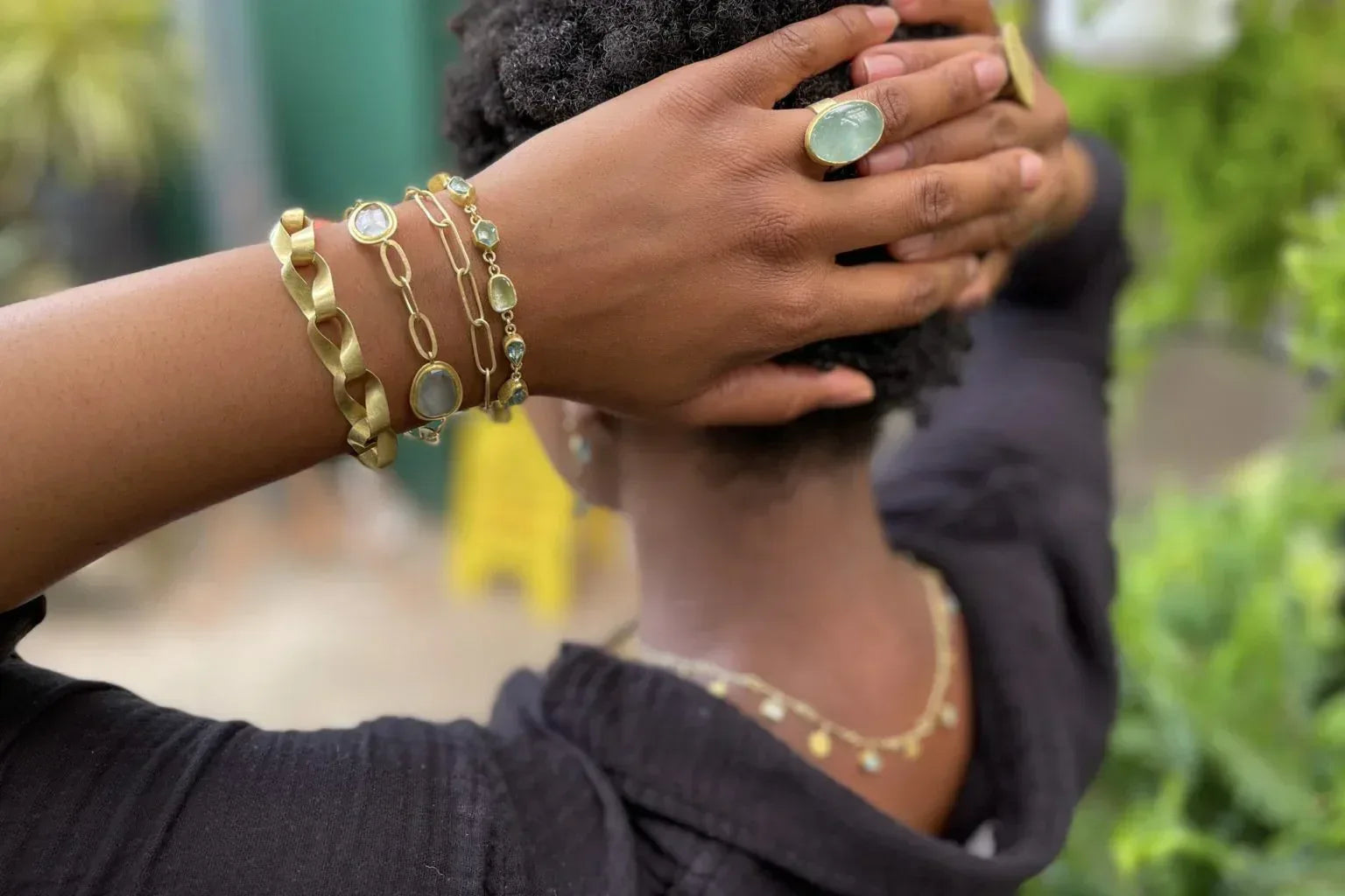 Person wearing gold bracelets and a ring with a blurred outdoor background