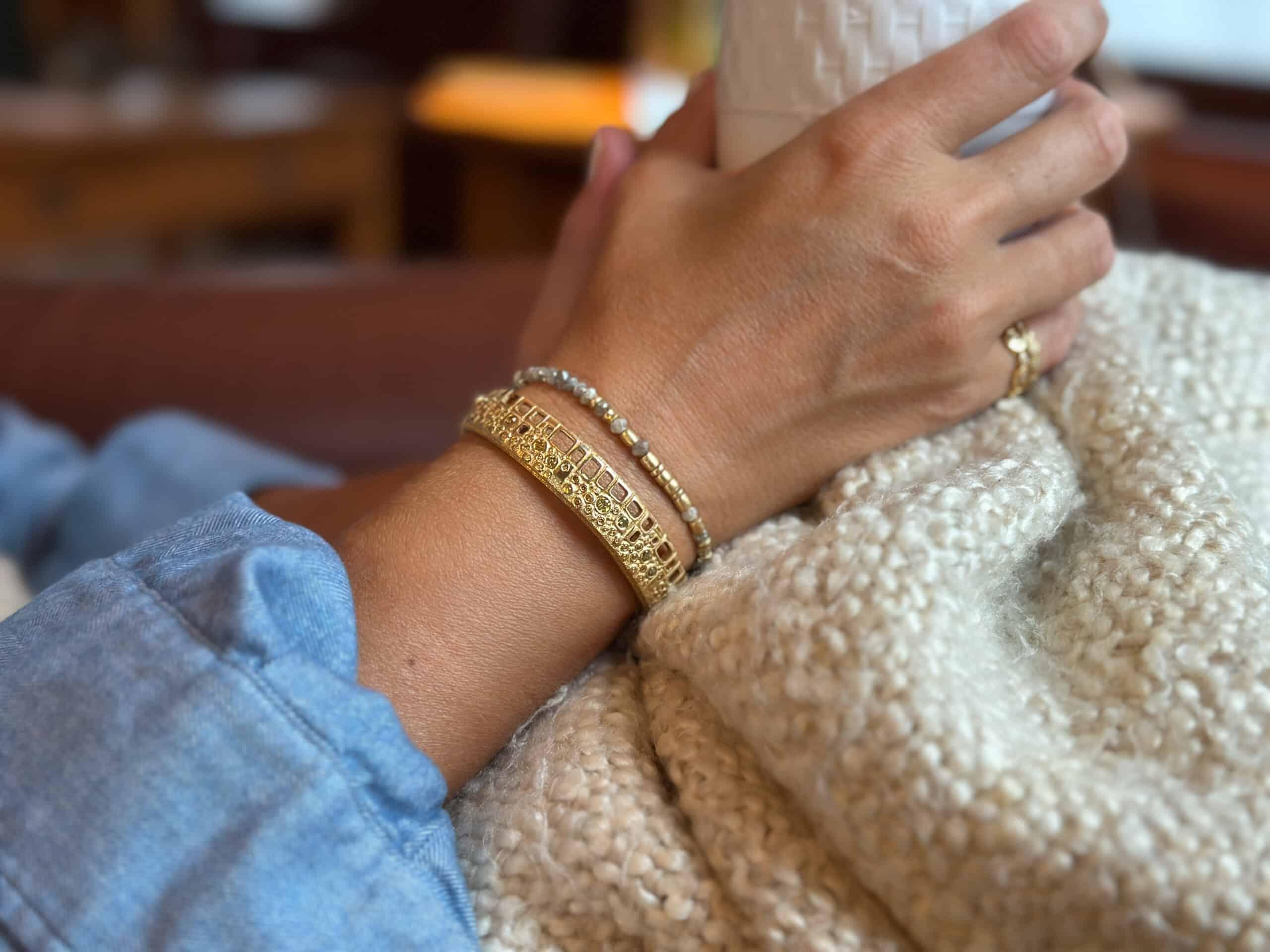 Close-up of a hand wearing gold bracelets and a ring, holding a white mug with a blurred background.