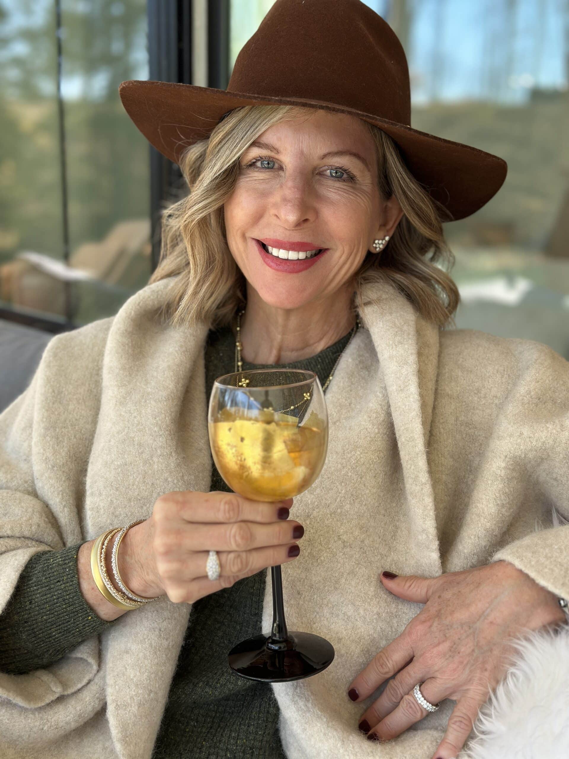 Woman in a beige coat and brown hat holding a glass of orange liquid, smiling.