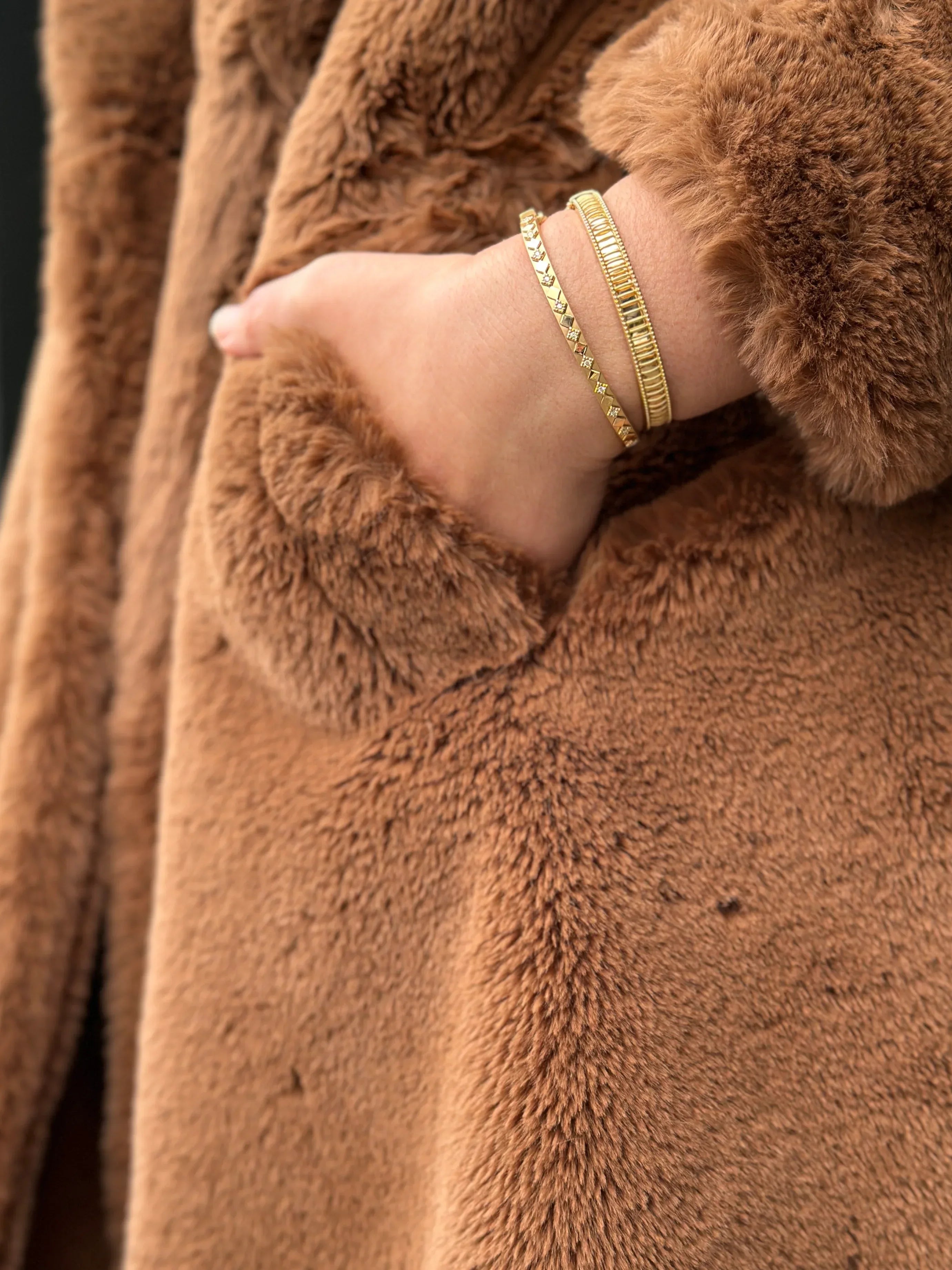 Close-up of a person wearing a brown teddy coat with gold bracelets on a blurred background