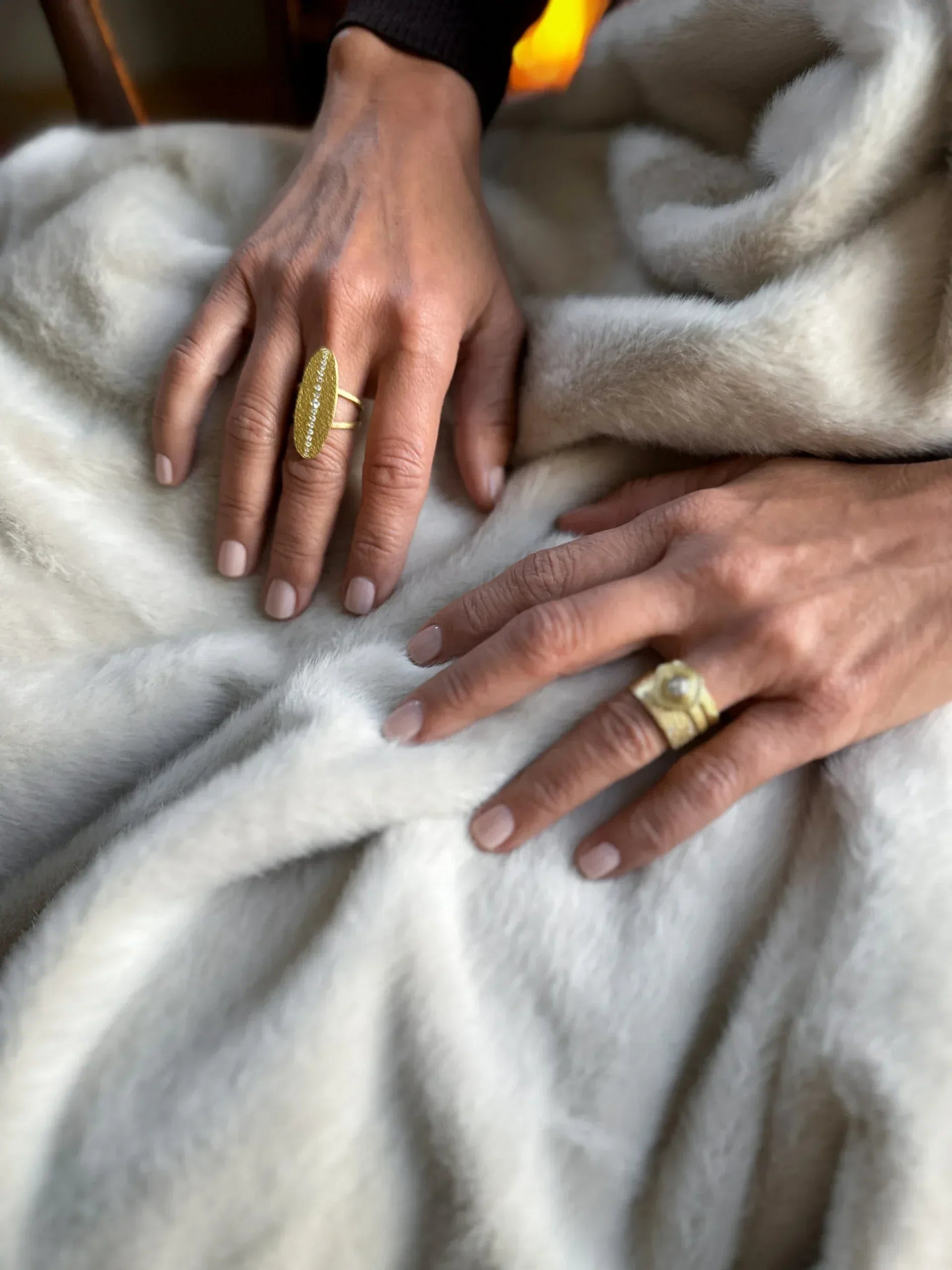Two hands with manicured nails rest on a soft, light faux fur blanket, each wearing the Yellow Gold and Diamond Oval Statement Ring by Petra Class—one oval-shaped and textured, the other featuring sparkling diamonds.