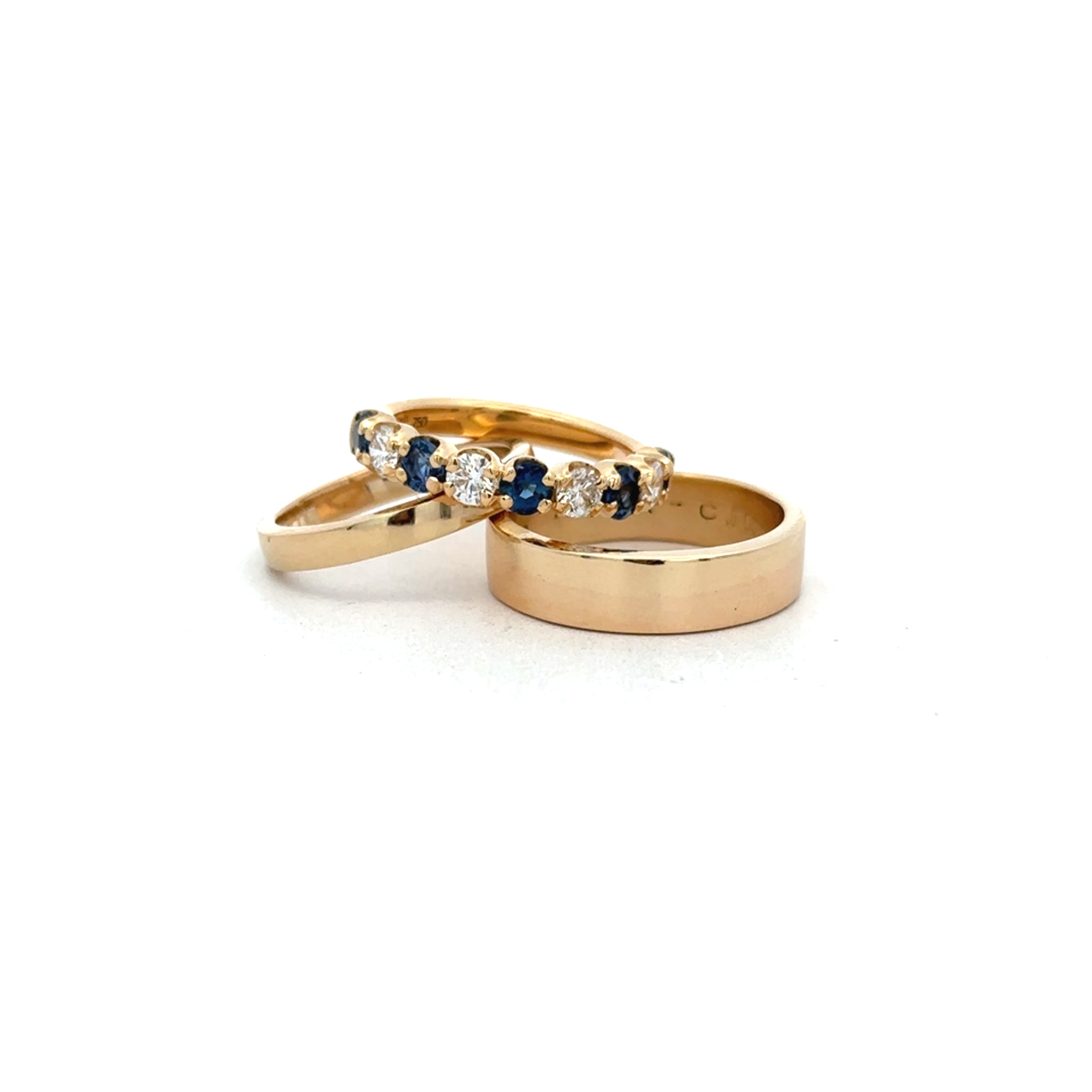Three gold rings on a white background, including a band ring and two stacked rings with small blue and clear gemstones.