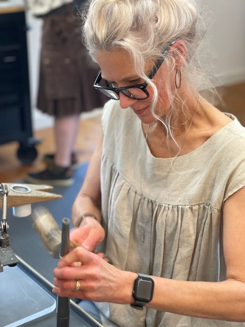 A woman with blonde hair, glasses, and a beige sleeveless top is shown from the waist up, working at a metalworking bench with a hammer and metal rod. Another person is blurred in the background.