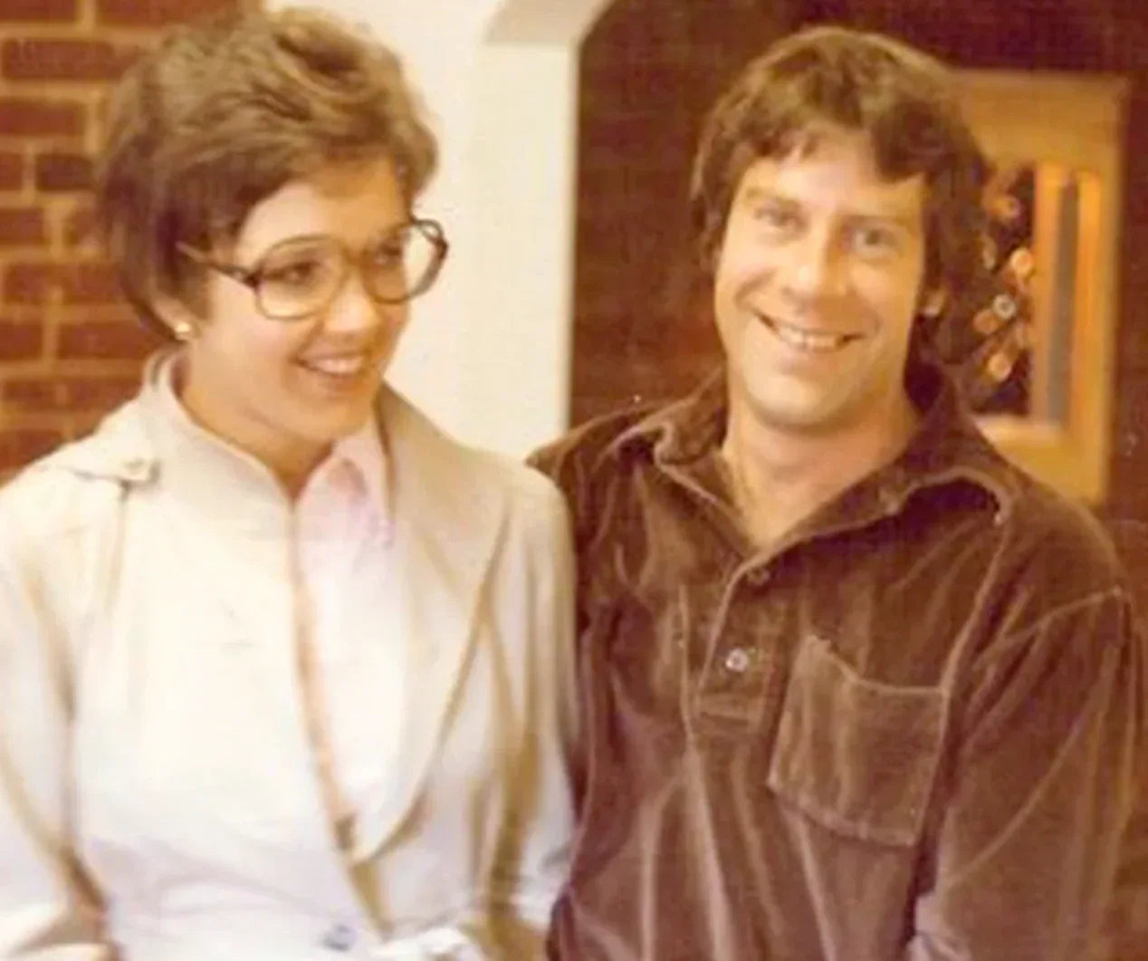 Two people standing close together indoors with a brick wall background