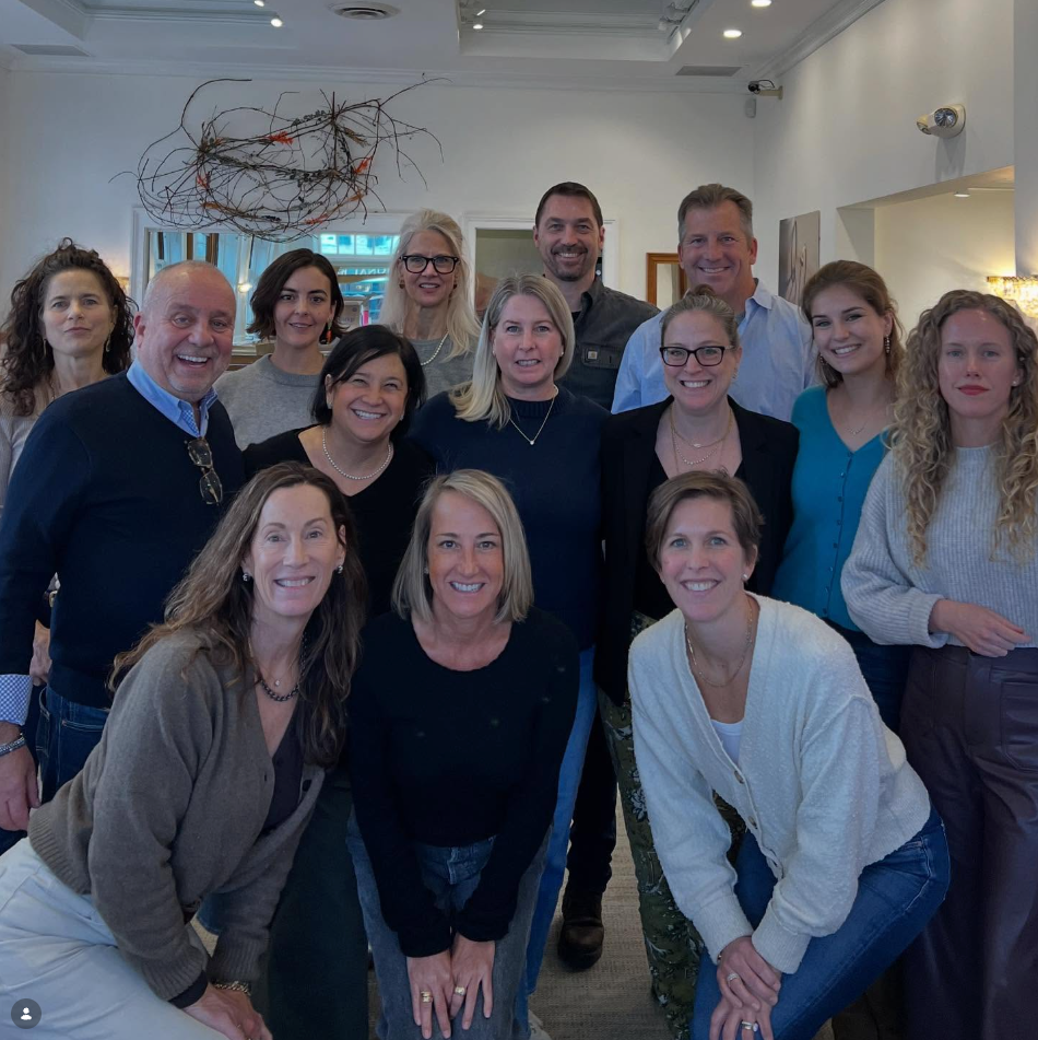 A group of 15 people smiling and posing together in a bright, modern indoor office or studio space with decorative wall art and warm lighting.