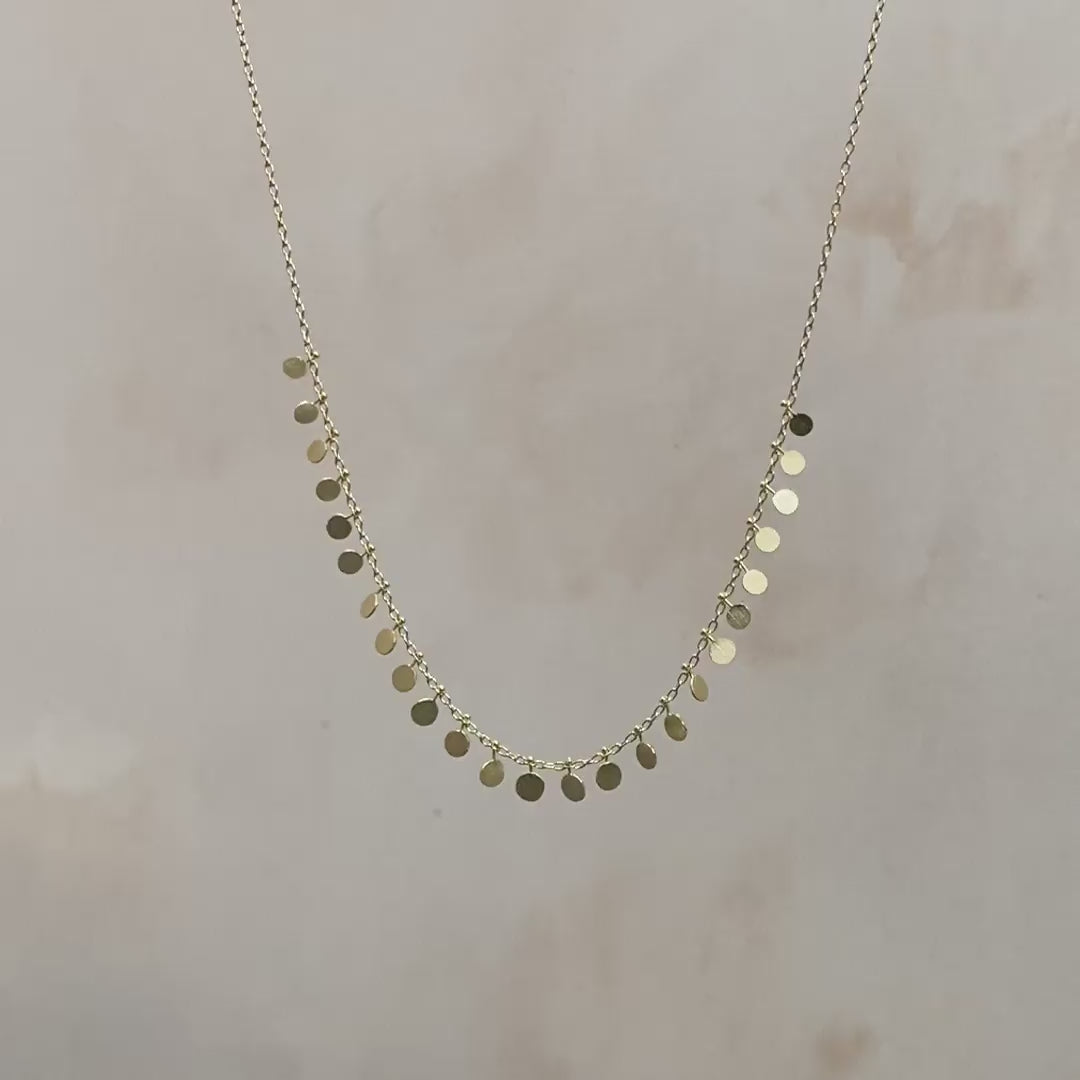 Video of a gold chain with gold dot pendants hanging from the chain and shimmering in the light on a grey background.