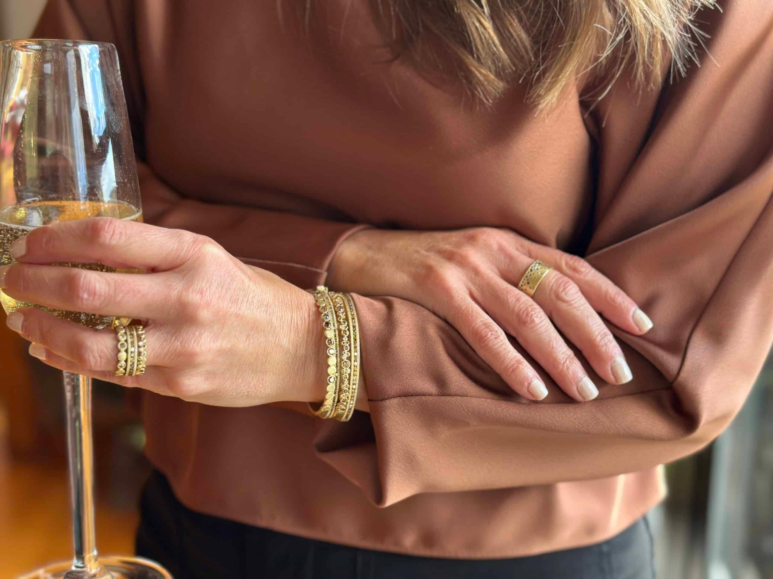 Person holding a glass of white wine with gold rings and bracelets on fingers.